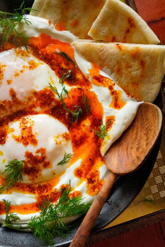 a side view of a plate of Cilbir (Turkish poached eggs) sitting on top of smooth Greek yogurt, bathed in an Aleppo garlic butter sauce, sprinkled with chopped fresh dill, and served with pita bread