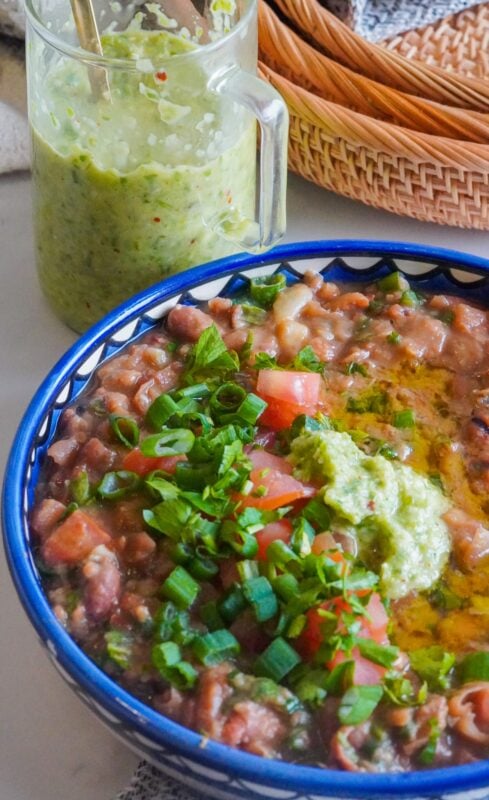 On a plain area, a wooden basket is placed near a glass jar that contains a silver spoon and a green spicy mixture. Past them, there is a plate of stewed dried fava beans with vegetables and oil on top.
