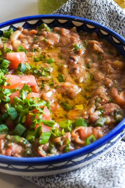 Part of a large plate is near a piece of cloth. Inside the plate, there's stewed fava beans or ful medames combined with chopped veggies and olive oil.