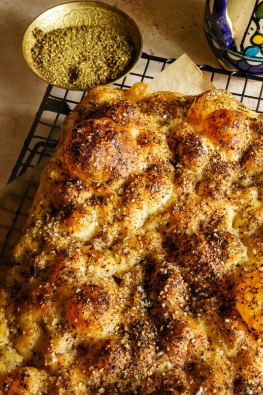 Crispy focaccia bread topped with zaatar and sumac placed on a wire rack, with a small bowl of zaatar on the side.