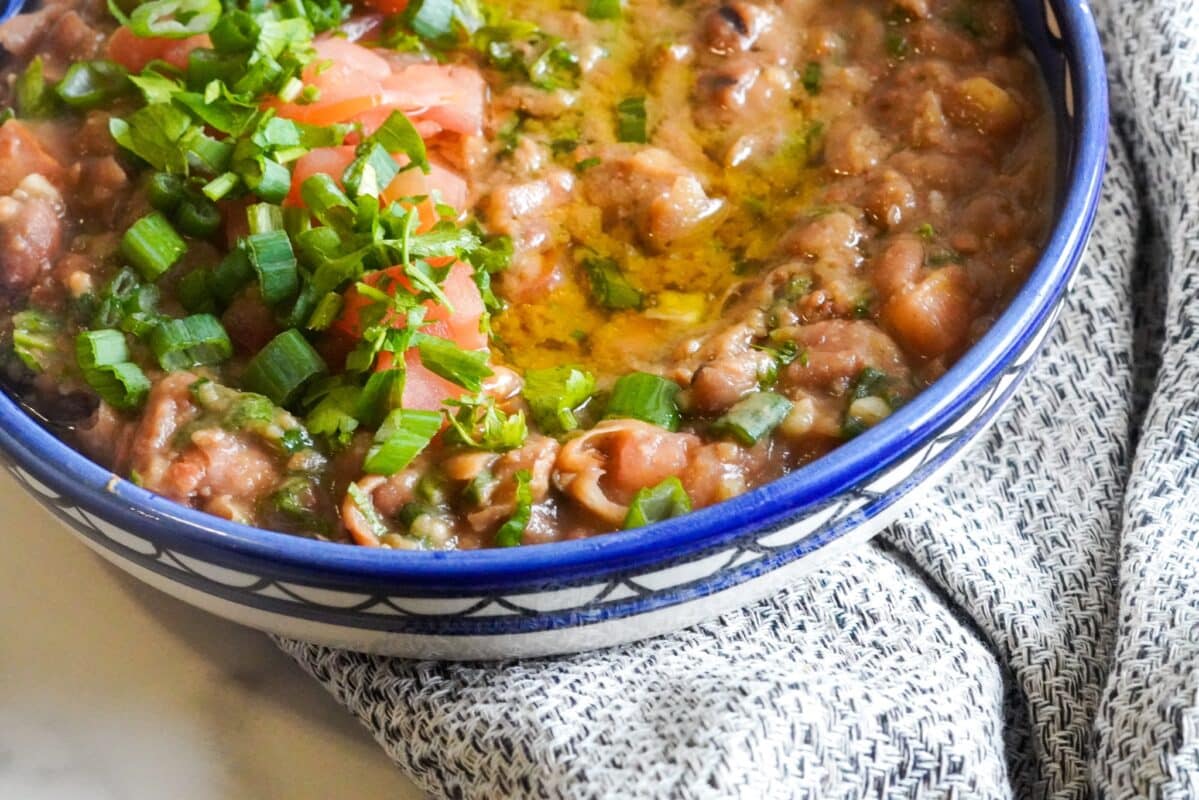 A piece of cloth holds a medium bowl. Inside the bowl, olive oil and some diced vegetables garnish a recipe of ful medames.