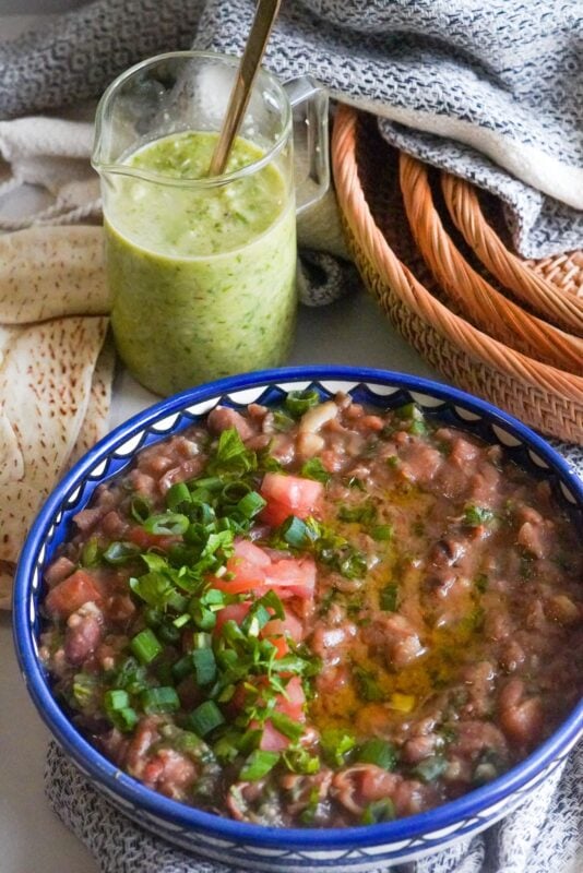 A table has a bamboo basket, a jar of green spicy sauce, and pita bread. On the table, there's also a dish of ful mudammas recipe garnished with chopped tomatoes, greens, and olive oil