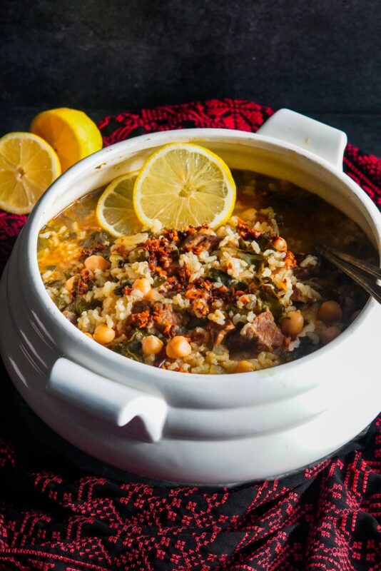 White porcelain soup pot filled with Gazan stew. It is made up of Swiss chard, beef chunks, rice, chickpeas, and topped with shatta. The pot is decorated with two slices of fresh lemon.