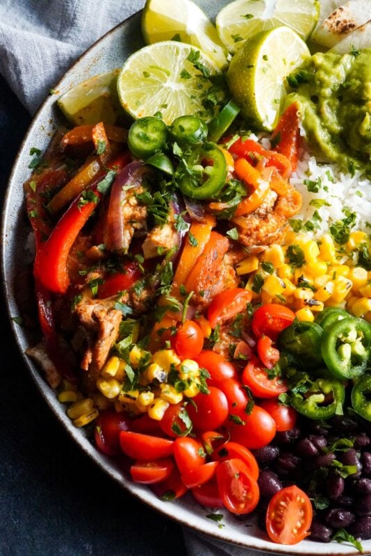 fajita with rice in a bowl topped with peppers, tomatoes, corn, black beans, guacamole, and slices of lemon