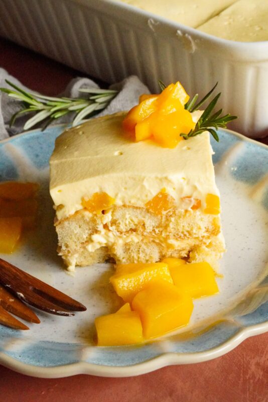 A delectable slice of Mango Tiramisu is placed on a plate. This tiramisu features layers of ladyfinger biscuits soaked in mango juice, topped with mango chunks and rosemary spring. 