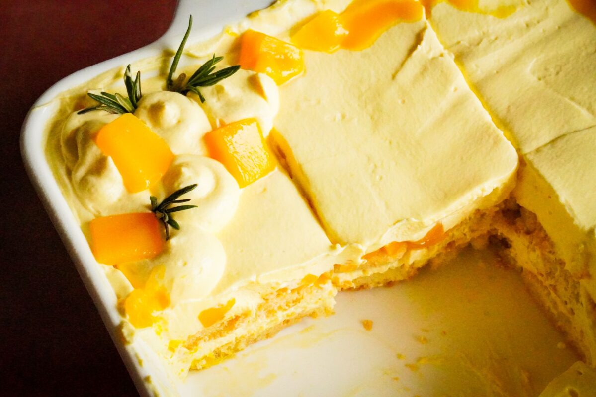 A pan of mango tiramisu that showcases some missed slices. This tiramisu is topped with mango chunks and rosemary sprigs.