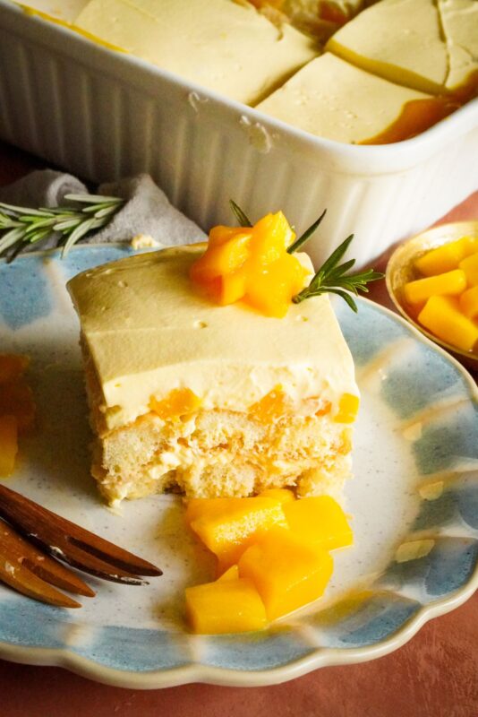 A slice of mango tiramisu is served placed on a plate and topped with mango chunks and rosemary spring. 