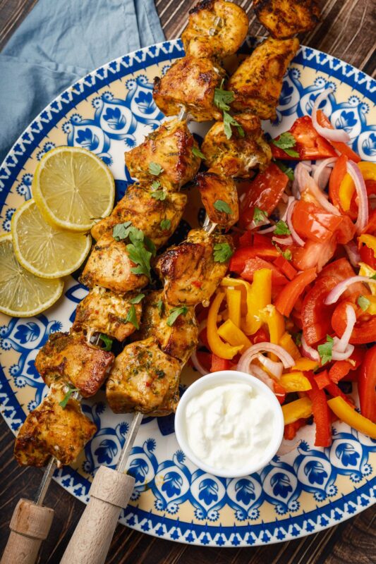 a plate of two skewers of Middle Eastern chicken kebabs sprinkled with finely chopped parsley served with Arabic chopped salad, a small bowl of garlic sauce, and lemon wedges