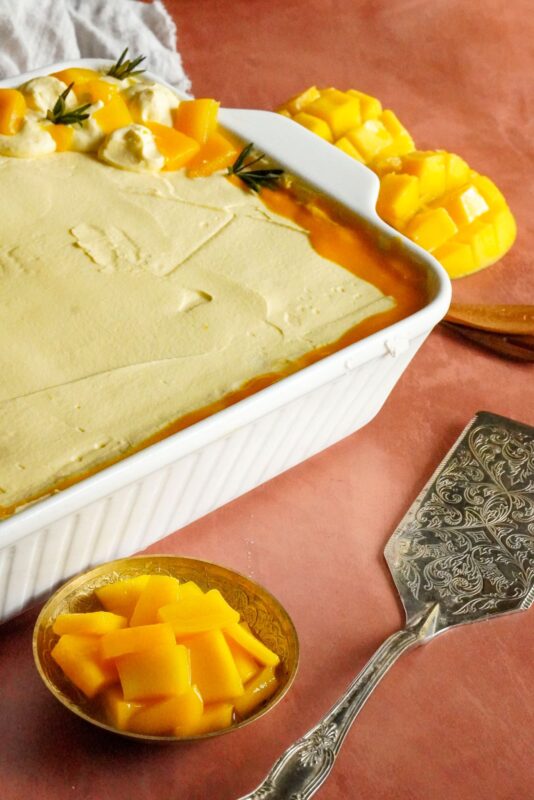 A pan of mango tiramisu dessert is topped with mango chunks and rosemary sprigs. The pan is placed beside a small bowl that contains mango chunks.