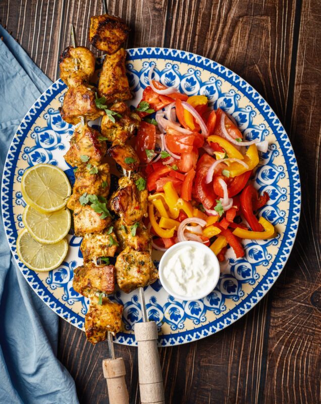 a top view of a plate of two skewers of shish tawook sprinkled with finely chopped parsley served with Arabic chopped salad, a small bowl of garlic sauce, and lemon wedges