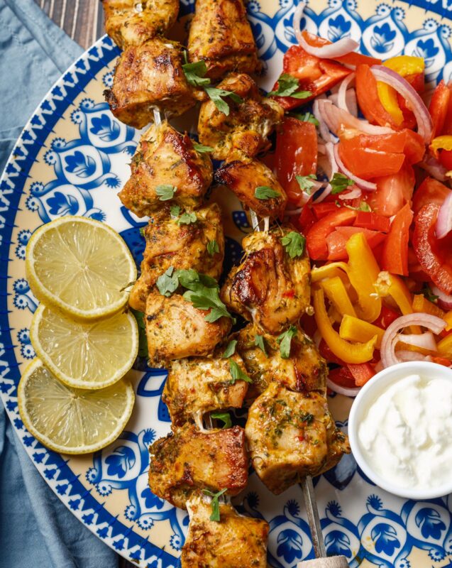 a close view of a plate of two skewers of shish tawook grilled chicken sprinkled with finely chopped parsley served with Arabic chopped salad, a small bowl of garlic sauce, and lemon wedges