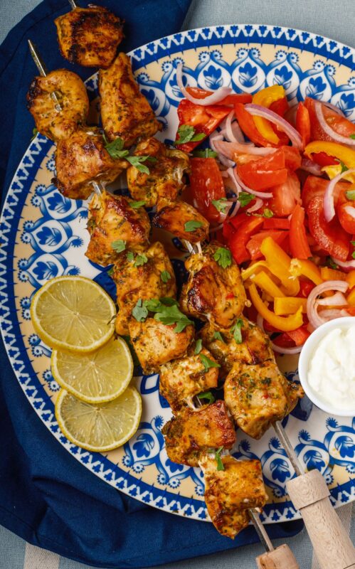 a plate of two skewers of Middle Eastern chicken shish tawook sprinkled with finely chopped parsley served with Arabic chopped salad, a small bowl of garlic sauce, and lemon wedges