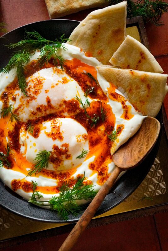 a top view of a plate of Cilbir (Turkish eggs) sitting on top of smooth Greek yogurt, bathed in an Aleppo garlic butter sauce, sprinkled with chopped fresh dill, and served with pita bread