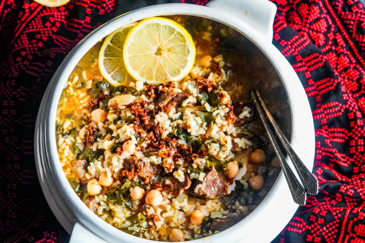 A white porcelain soup pot filled with Fogaiyya stew, which is made up of chickpeas, beef chunks, cooked rice, and Swiss chard topped with Palestinian shatta, and is decorated with two slices of fresh lemon.