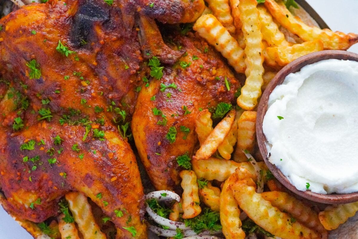 Image of sumac-spiced oven-baked chicken on a bed of onions, accompanied by crinkle-cut fries, and a bowl of creamy garlic paste.