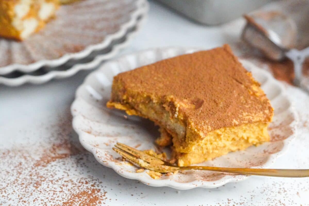 A square piece of pumpkin tiramisu garnished with ground cinnamon and cocoa is set on a plate with a sea-shell shape. The piece is partly eaten.