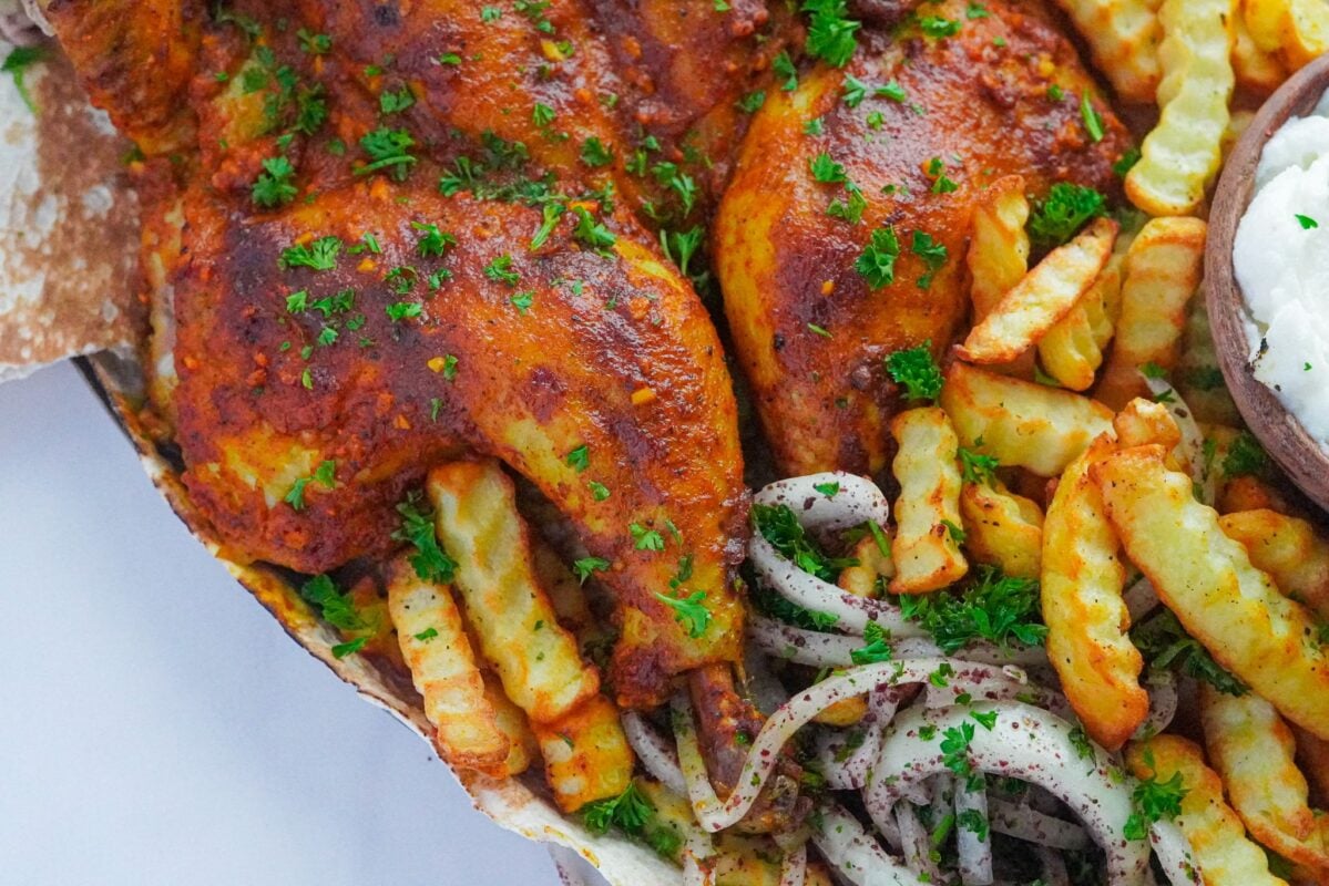 A baked chicken with brown crust garnished with parsley, with French Fries, Chopped onions and Sumac, and garlic paste on the side.