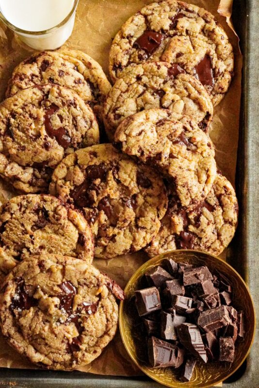 A tray packed with delicious brown butter chocolate chip cookies topped with sea salt, with some chocolate chunks and a glass of milk on the side.