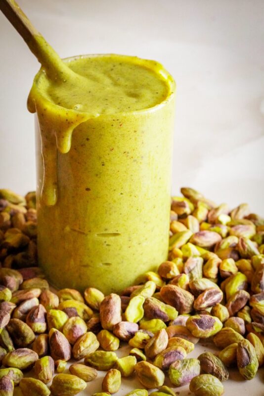 A tall glass filled to the brim with green pistachio spread sits amidst a bunch of pistachios, with the spread dripping down its sides