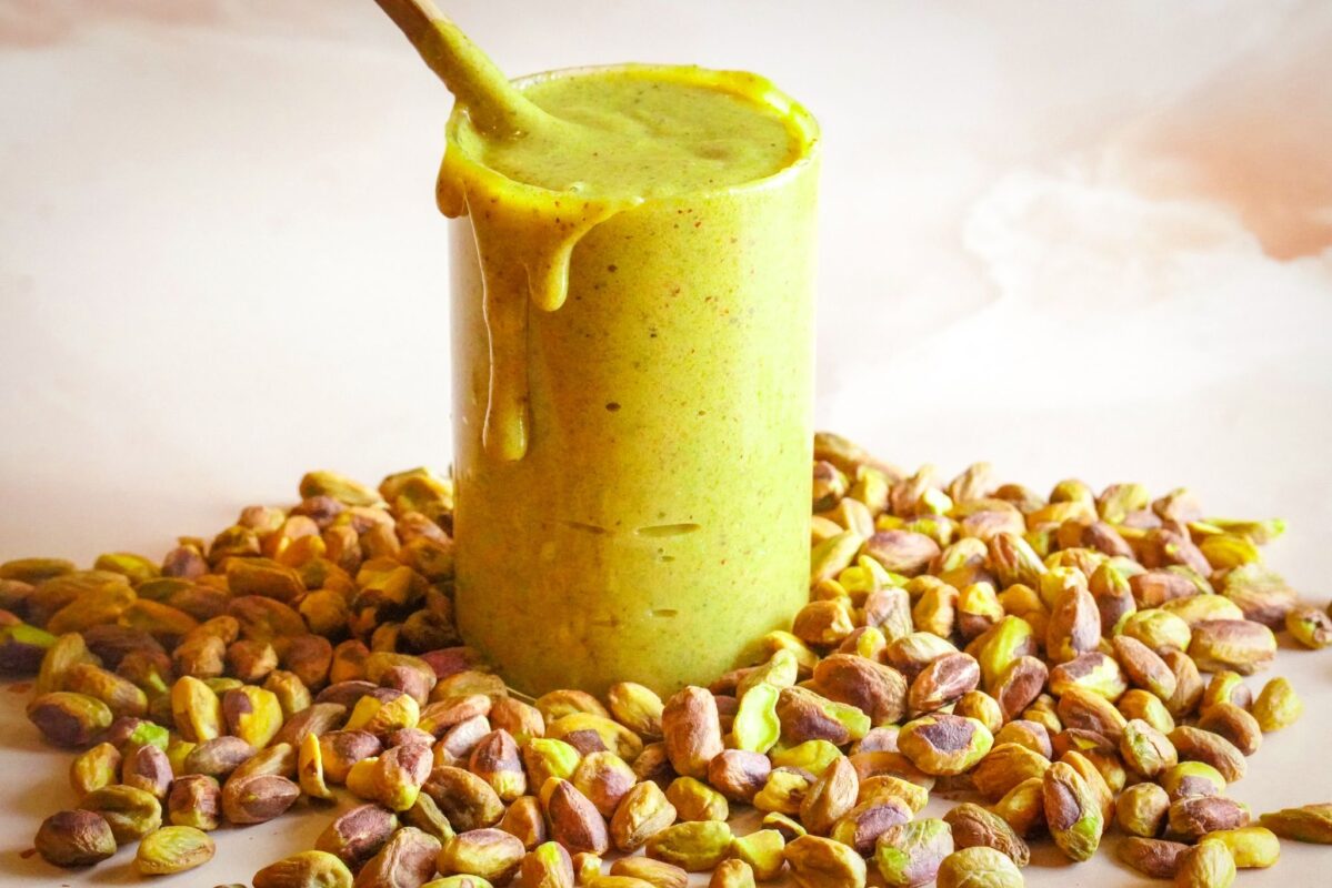 A tall glass filled to the brim with green pistachio spread sits amidst a bunch of pistachios, with the spread dripping down its sides.