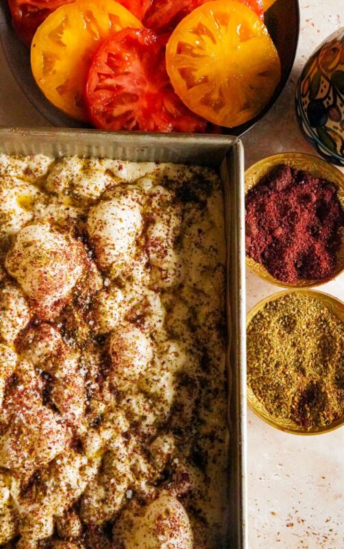 Raw, airy and bubbly focaccia dough placed in a baking pan, topped with a combination of zaatar, sumac, and olive oil. Three bowls placed on the side, one for the sumac and the other for the zaatar, and the third for the red and yellow thinly sliced tomatoes.