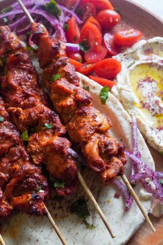 Three chicken skewers are placed on top of pita bread garnished with parsley with some cherry tomatoes and onion rings.
