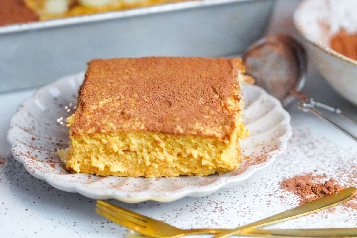 A square piece of pumpkin tiramisu is set on a plate with a sea-shell shape. It is garnished with ground cinnamon and cocoa.