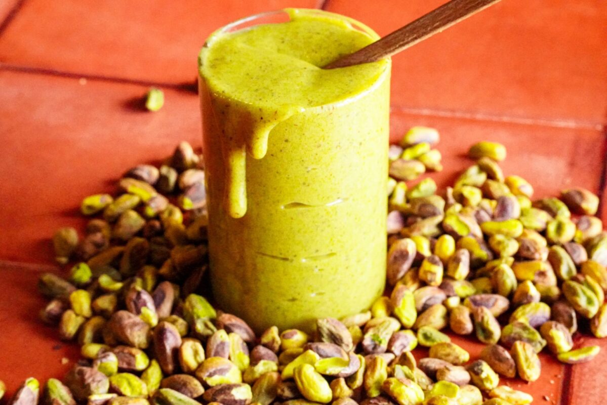 A tall glass filled to the brim with green pistachio spread sits amidst a bunch of pistachios, with the spread dripping down on one side.