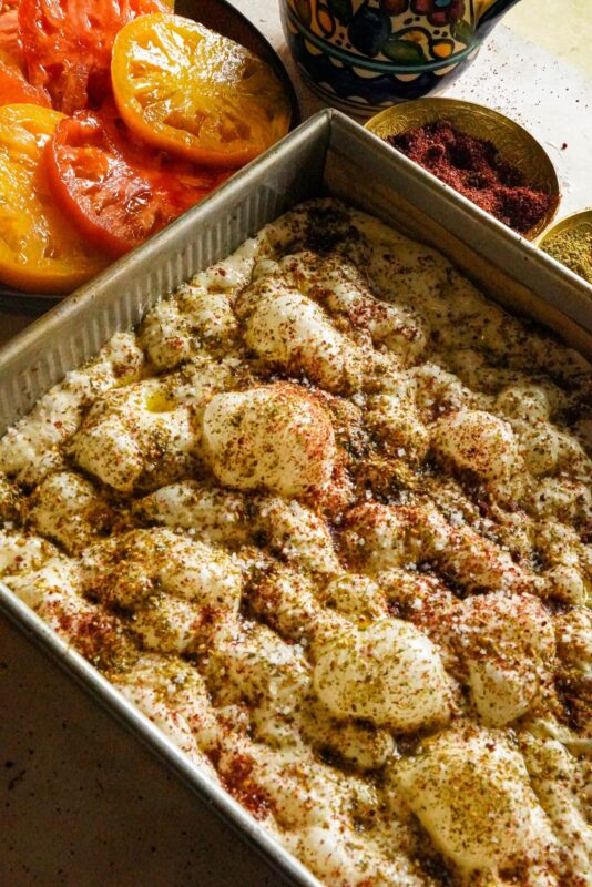 Raw, airy and bubbly focaccia dough placed in a baking pan, topped with a combination of zaatar, sumac, and olive oil. Three bowls placed on the side, one for the sumac and the other for the zaatar, and the third for the red and yellow thinly sliced tomatoes.