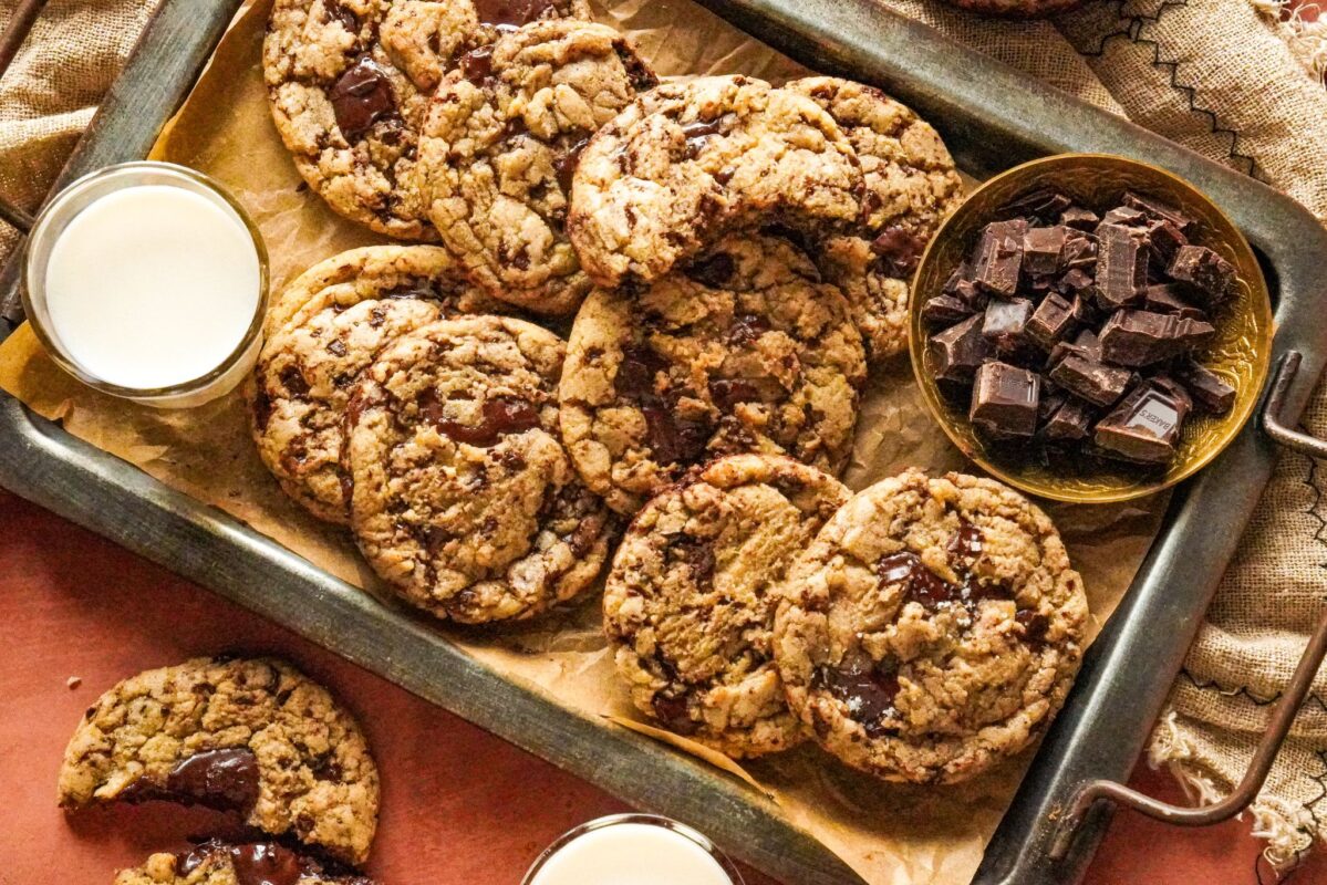 A tray packed with irresistible brown butter chocolate chip cookies with two glasses of milk and some chocolate chunks on the side. The cookies are topped with sea salt.