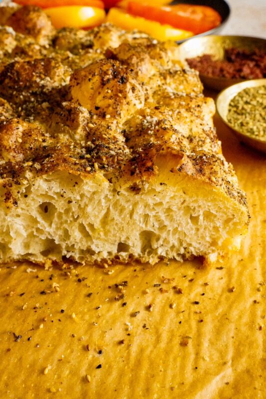 A fluffy piece of focaccia bread topped with sumac and zaatar, with some thinly sliced red and yellow tomatoes in the background.