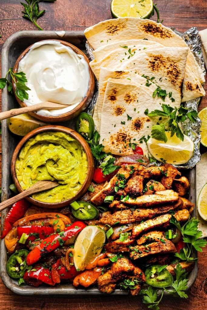 top shot of a sheet pan of tender juicy chicken served next to roasted bell pepper and onions, a bowl of sour cream, a bowl of guacamole, lemon wedges, and tortilla bread