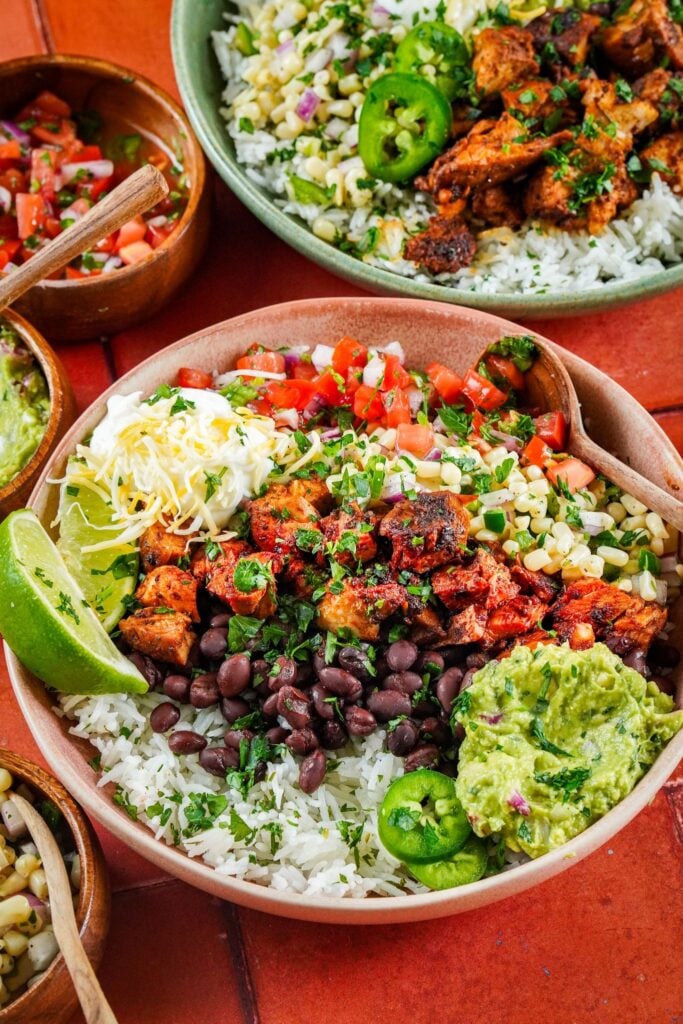 two bowls of chipotle chicken served on a bed of cilantro lime rice and topped with guacamole sauce, corn salsa, black beans, chopped tomatoes, lime wedges, and chopped fresh cilantro