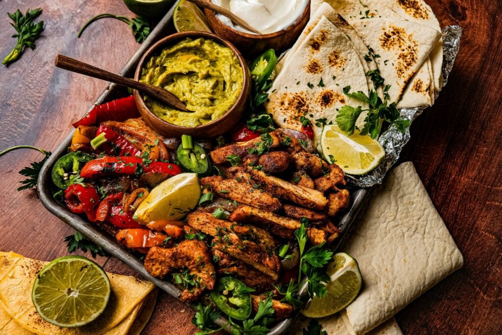 a sheet pan of tender juicy chicken fajitas served next to roasted bell pepper and onions, a bowl of guacamole, lemon wedges, and tortilla bread