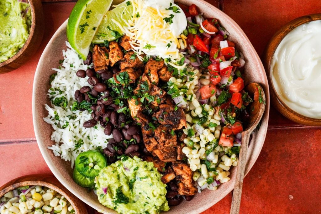 a top shot of copycat chipotle chicken served in a bowl on a bed of cilantro lime rice and topped with guacamole sauce, corn salsa, black beans, lime wedges, and chopped fresh cilantro