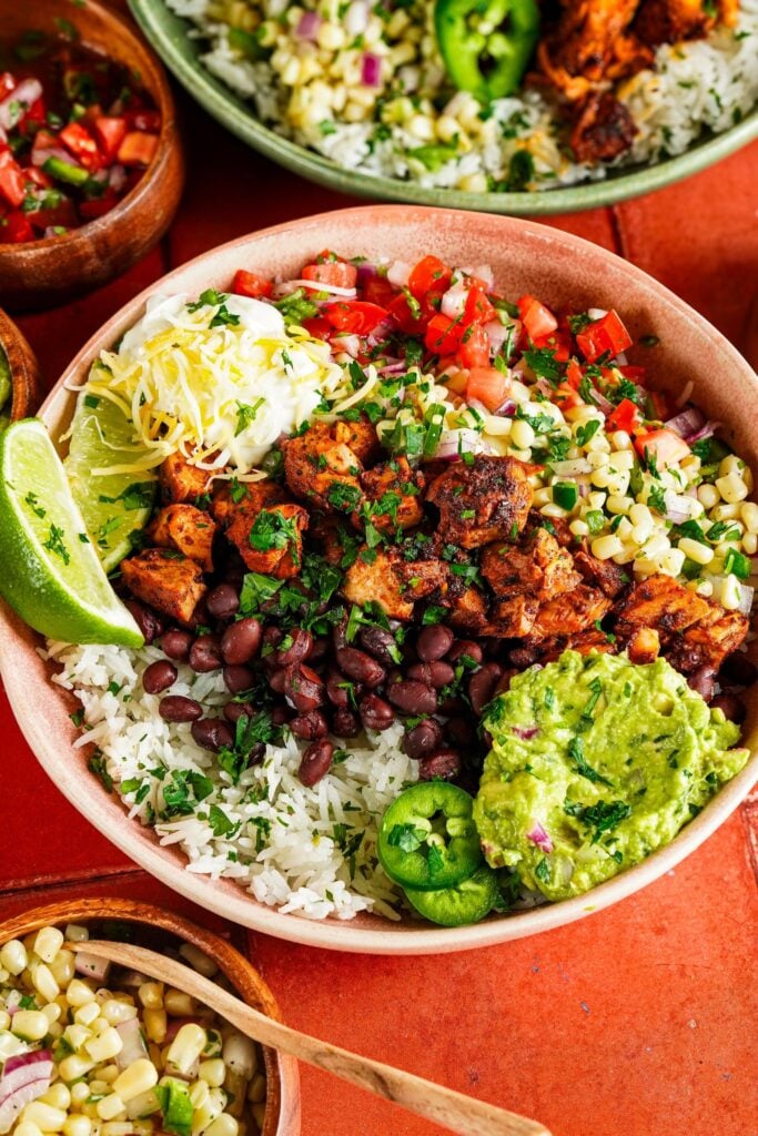 chipotle chicken served in a bowl on a bed of cilantro lime rice and topped with guacamole sauce, corn salsa, black beans, chopped tomatoes, lime wedges, and chopped fresh cilantro