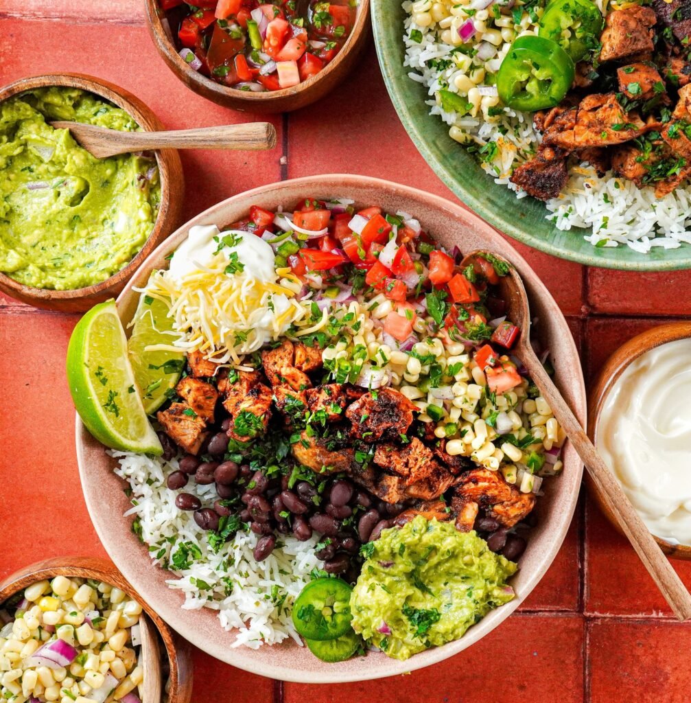 a top shot of chipotle burrito bowl made with juicy chicken served on cilantro lime rice and topped with guacamole sauce, corn salsa, black beans, lime wedges, and chopped fresh cilantro served next to a bowl of guacamole sauce and another bowl of corn salsa