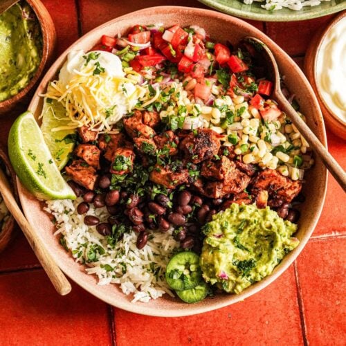 a top shot of copycat chipotle chicken burrito bowl is served on a bed of cilantro lime rice and topped with guacamole sauce, corn salsa, black beans, lime wedges, and chopped fresh cilantro