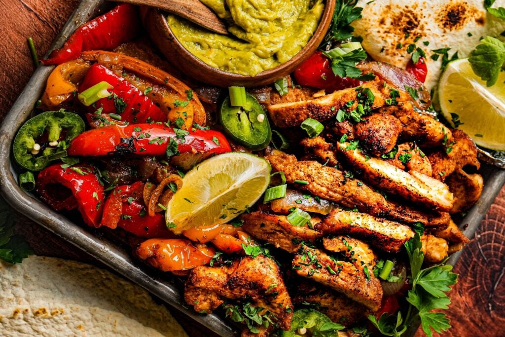 close up shot of tender juicy chicken fajitas served on a sheet pan next to roasted bell pepper and onions, a bowl of guacamole, and lemon wedges