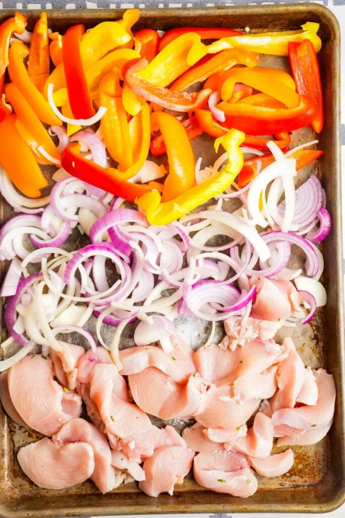 a sheet pan of raw chicken breasts, uncooked red and yellow bell pepper, and uncooked red and yellow onion