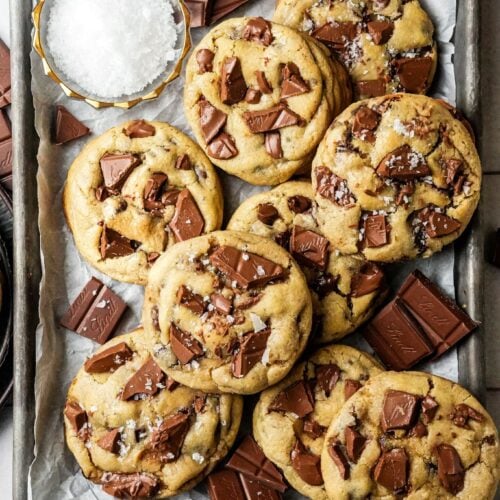 top shot of Levain Bakery cookies, topped with extra chocolate chips and a sprinkle of flaky sea salt, are placed on a baking sheet next to a bowl of flaky sea salt