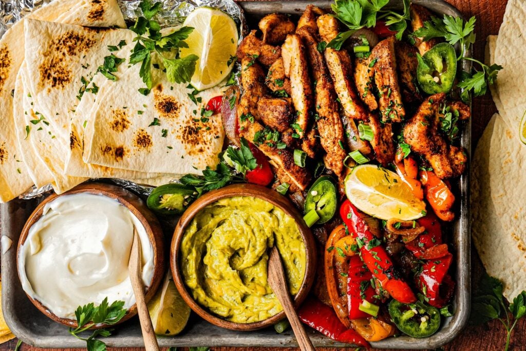 a sheet pan of tender juicy chicken fajitas served next to roasted bell pepper and onions, a bowl of sour cream, a bowl of guacamole, lemon wedges, and tortilla bread