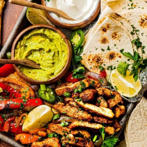sheet pan of tender juicy chicken fajitas served next to roasted bell pepper and onions, a bowl of sour cream, a bowl of guacamole, lemon wedges, and tortilla bread