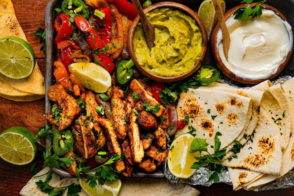 a sheet pan of tender juicy chicken fajitas served next to roasted bell pepper and onions, a bowl of sour cream, a bowl of guacamole, lemon wedges, and tortilla bread