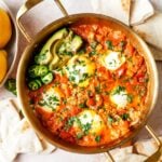 top shot of a skillet filled with shakshuka, garnished with avocado slices, jalapeños, and fresh parsley, surrounded by pita bread and a bowl of lemons