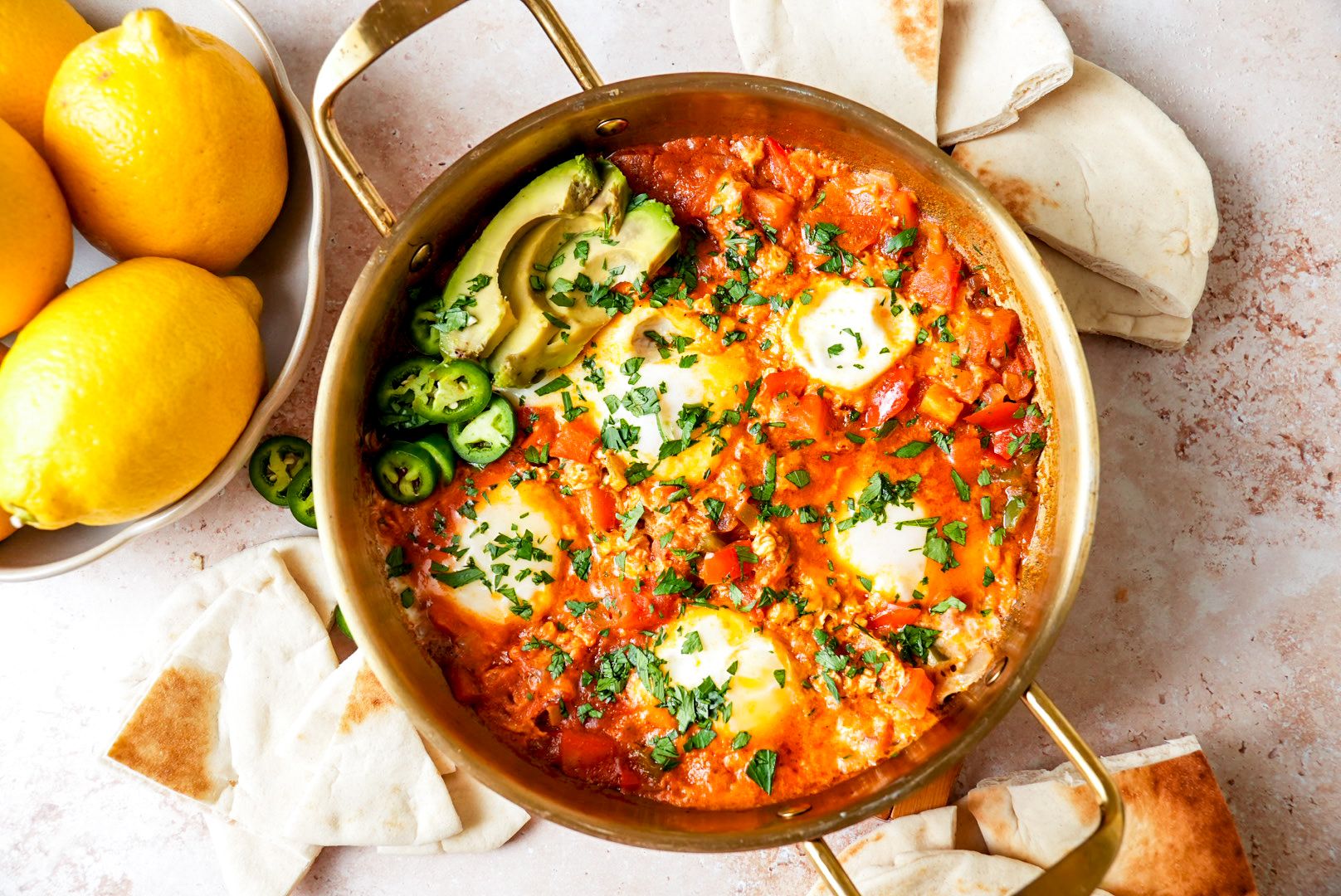 top shot of a skillet filled with shakshuka, garnished with avocado slices, jalapeños, and fresh parsley, surrounded by pita bread and a bowl of lemons