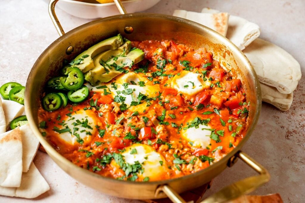 A pan of freshly made shakshuka with poached eggs in a rich tomato sauce, garnished with fresh parsley, sliced avocado, and jalapeños, served with pita bread