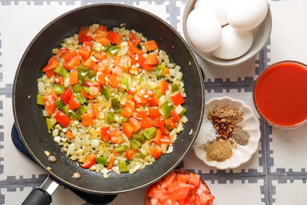 a pan of sauteed minced garlic, chopped onions, and diced bell pepper placed on low heat next to a bowl of tomato sauce, a plate of spices, and eggs