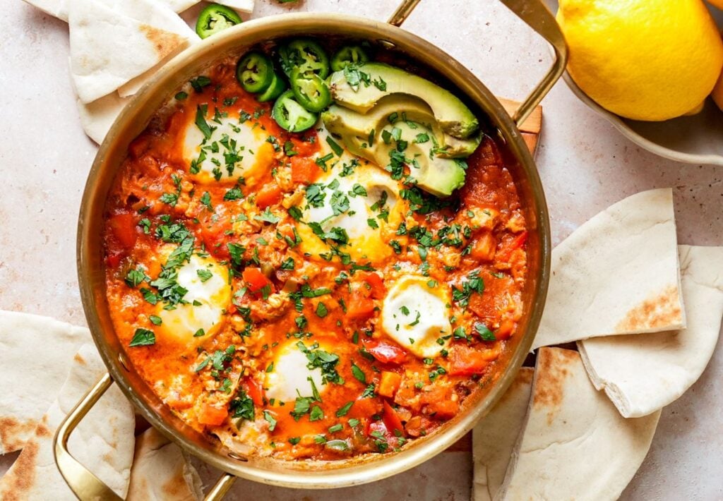 A pan of shakshuka garnished with fresh parsley, sliced avocado, and jalapeños, served with warm pita bread on the side