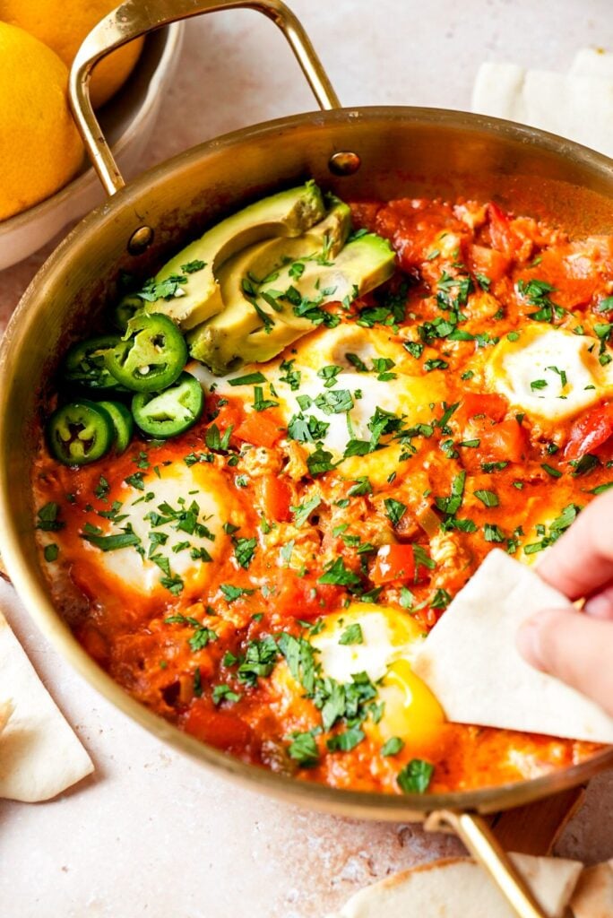 A woman scooping steaming shakshuka with pita bread from a skillet